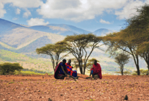 Maasai men in traditional clothing sitting under acacia trees with mountains in the background, Tanzania