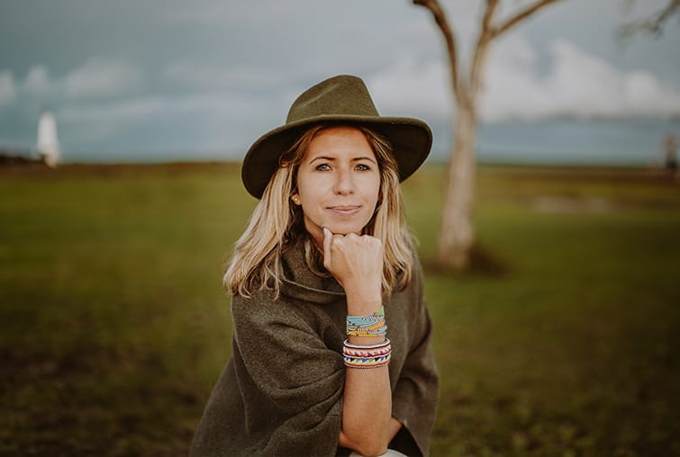 Woman wearing a green hat and poncho sitting outdoors on grassland with colourful beaded bracelets