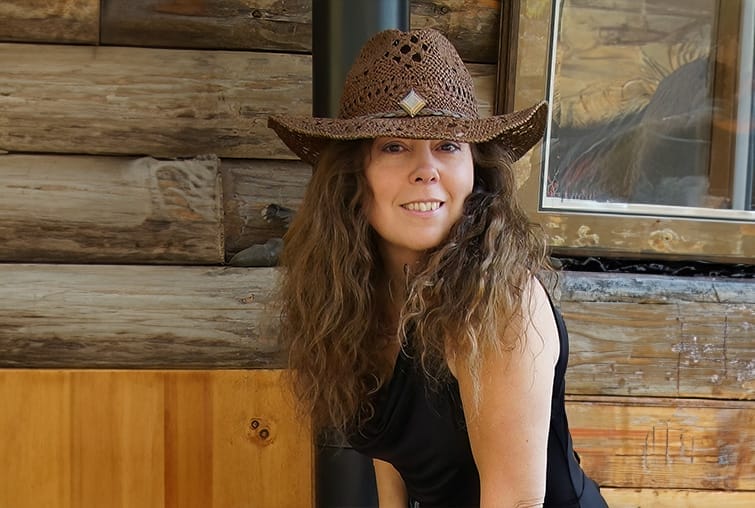 Smiling woman in a woven cowboy hat posing in front of rustic wooden cabin wall