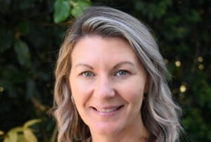 Smiling woman with blonde hair standing outdoors against a leafy green background