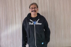 Smiling man with glasses and moustache wearing a black Trailblazer hoodie standing indoors
