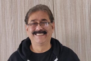 Smiling man with glasses and mustache wearing a black hoodie against wooden background