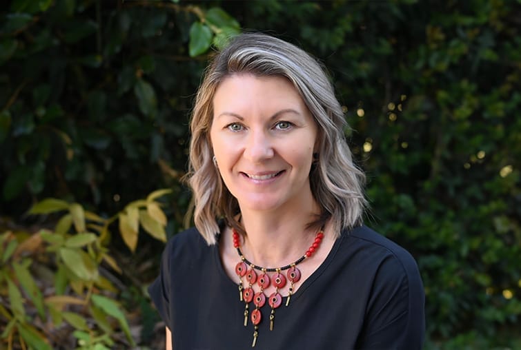 Smiling woman with blonde hair wearing a black top and red beaded necklace outdoors