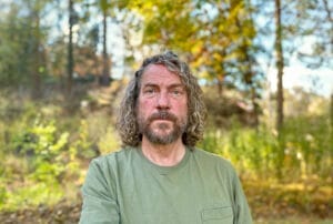 Man with curly hair and beard wearing a green shirt standing outdoors in a forest during autumn