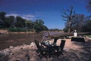 Riverside bush dining setup with tables and chef in safari wilderness