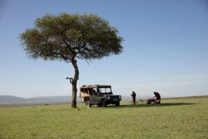 Safari jeep parked under acacia tree with guides setting up picnic on African savanna
