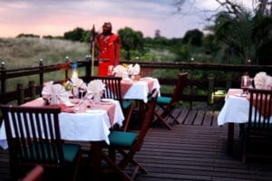 Romantic outdoor dining setup on safari lodge deck with Maasai host at sunset in Kenya