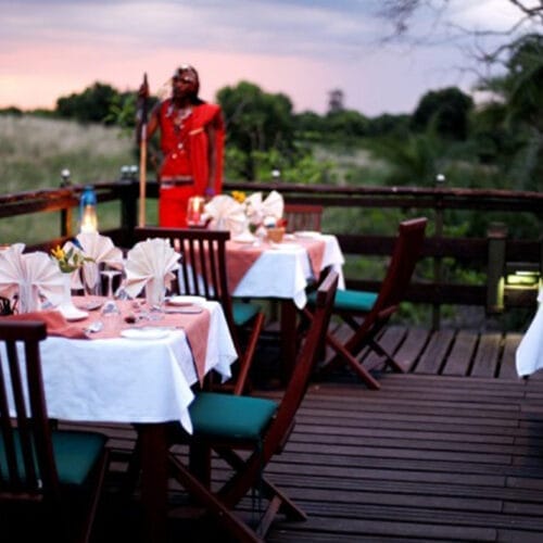 Untitled-3 | Gamewatchers Safaris Romantic outdoor dining setup on safari lodge deck with Maasai host at sunset in Kenya
