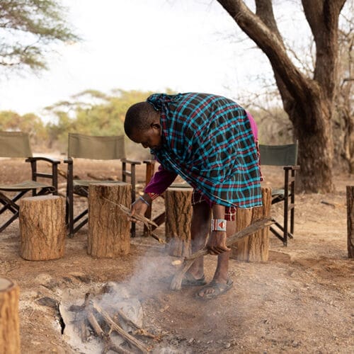 Maasai tribesman preparing campfire surrounded by wooden stools and chairs in safari camp
