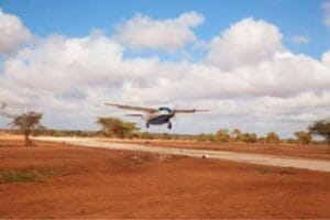 Small safari plane landing on dirt airstrip in Kenya with blue sky and scattered clouds