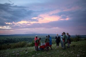 Tourists and Maasai guides enjoying sundowner drinks on safari in Kenya with scenic sunset view