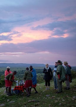 Tourists and Maasai guides enjoying sundowner drinks on safari in Kenya with scenic sunset view