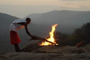 Person tending to a campfire on a rocky hilltop at sunset with mountain views