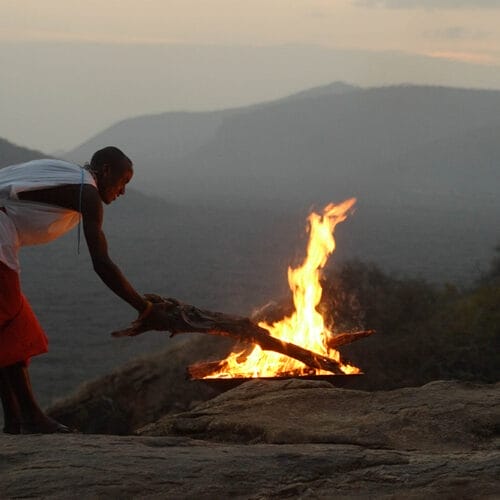 samburustaff | Gamewatchers Safaris Person tending to a campfire on a rocky hilltop at sunset with mountain views