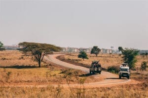 Two safari vehicles driving along a winding dirt road through dry grassland with scattered trees and city buildings in the distance