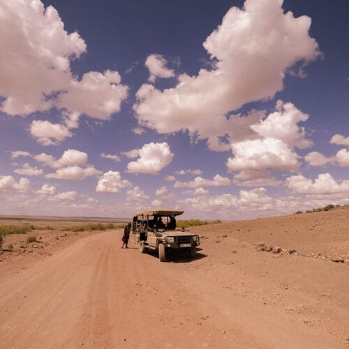 gallery3 | Gamewatchers Safaris Safari vehicle on a dusty road under a bright blue sky filled with scattered white clouds in a dry open landscape