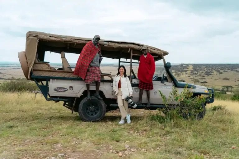 A lady holding professional telephoto lens with two Massai safari guides stating by safari vehicle at in Ol Kinyei Conservancy in Kenya. Photo credit Celine & Sacha