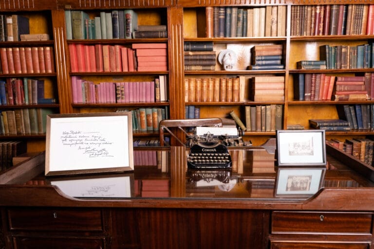 Vintage wooden desk with a black Corona typewriter, framed handwritten letter, and framed photo on a glass surface; tall wooden bookshelves in the background.
