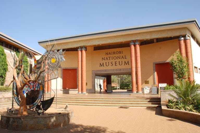 Entrance to Nairobi National Museum with a wide walkway and red pillars; a colorful sculpture stands to the left.