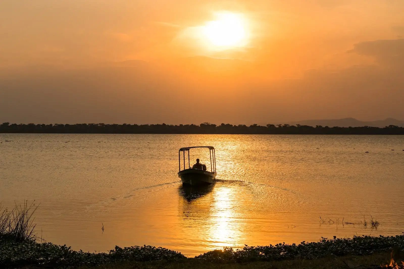 Silhouette of a small boat with a person on a calm lake at sunset, orange sky reflecting on the water at Honey Bear BUsh Camp, Uganda