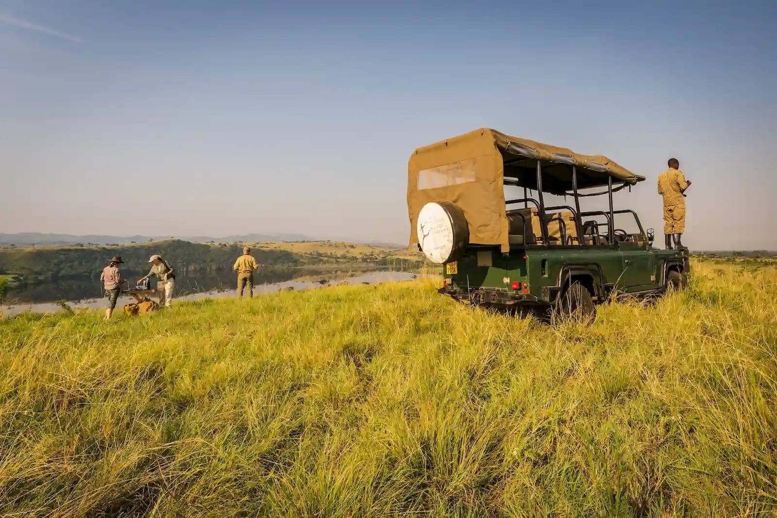 Green safari vehicle parked on tall grass, overlooking a river valley; a guide stands on the rear as others set up nearby ay Honey Bear Bush Camp, Uganda.