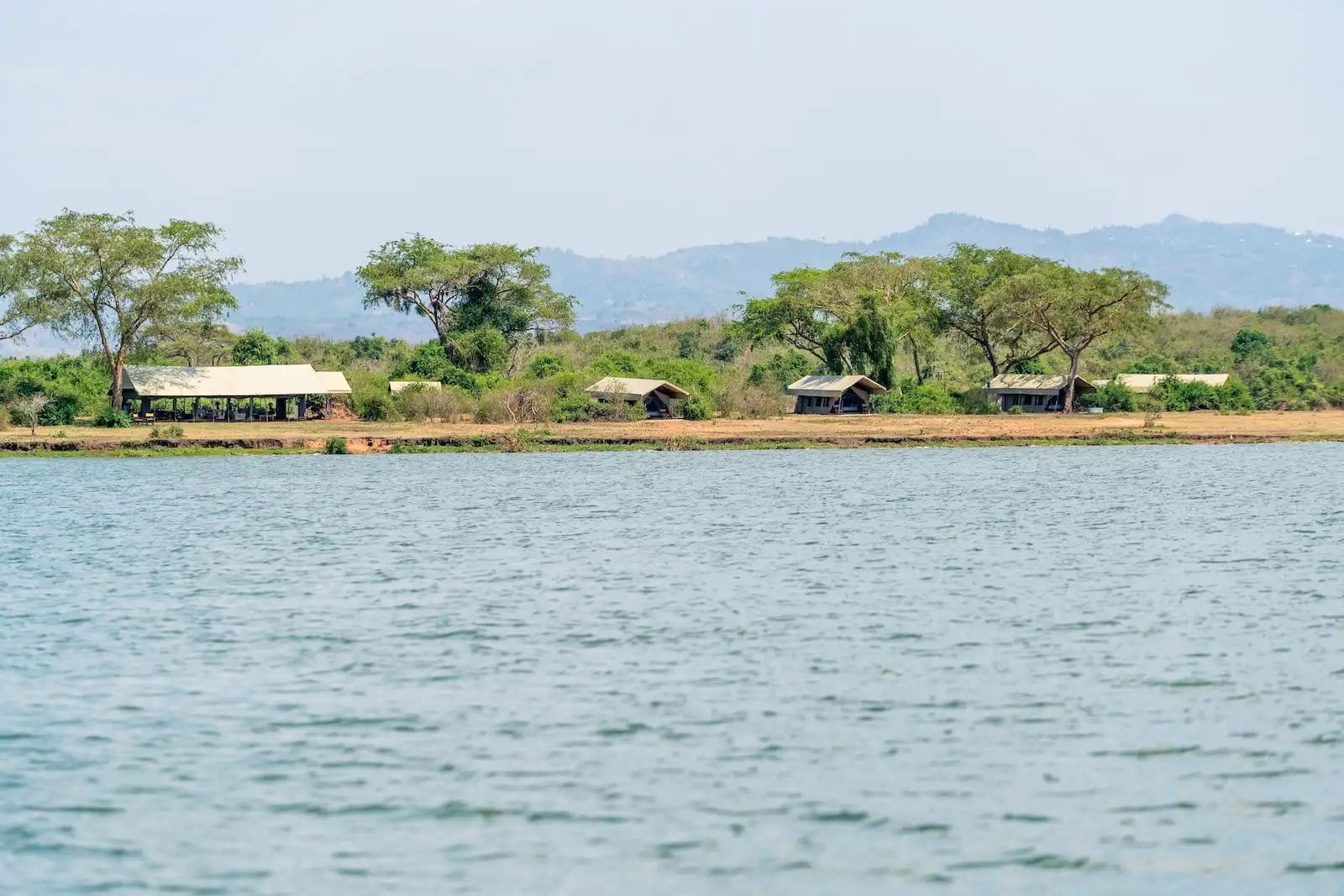 Lakeside camp with small thatched-roof huts and trees along the shore, hills in the distance. Honey Bear Bush Camp, Uganda.