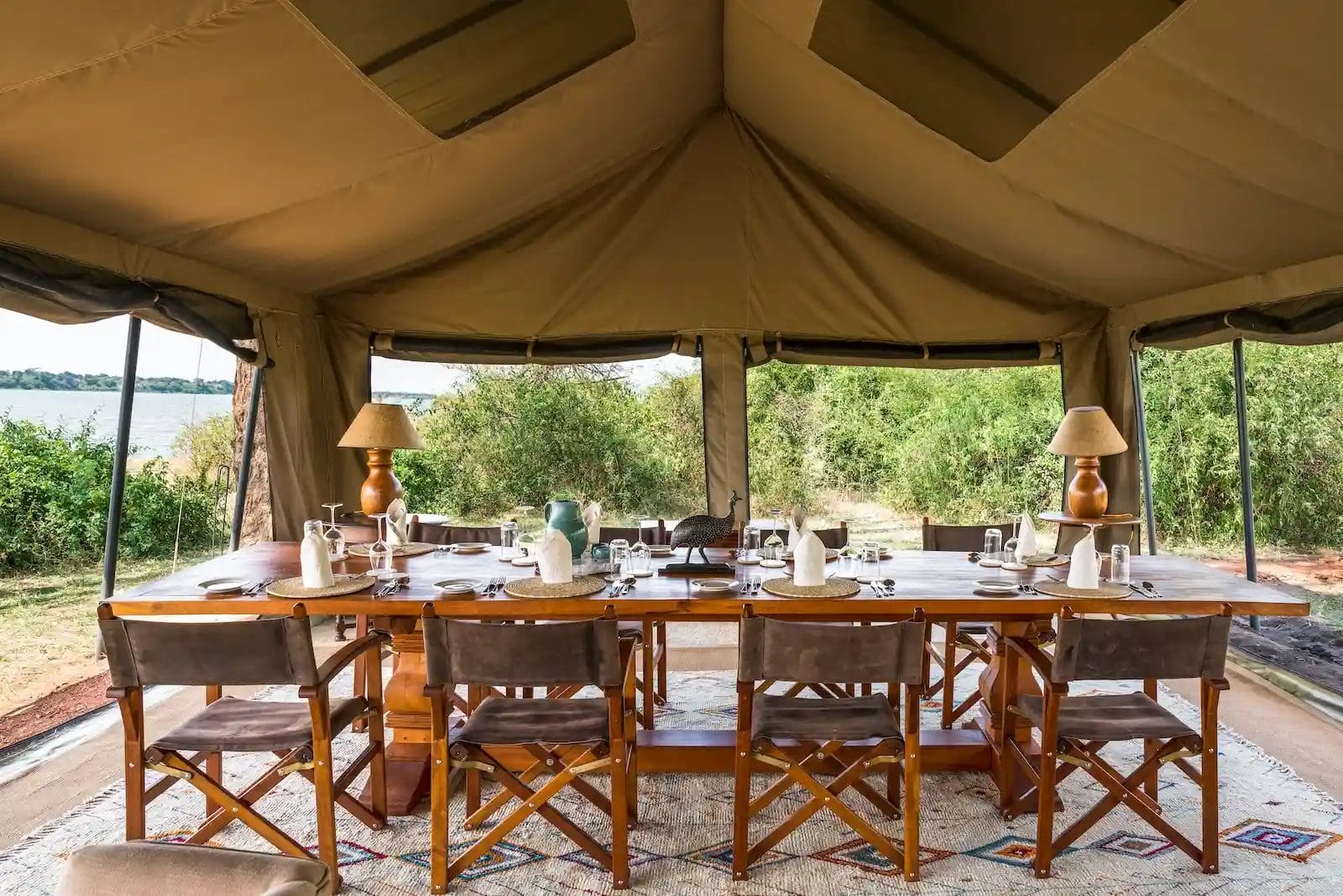 Outdoor safari-style dining setup inside a large beige canvas tent with a long wooden table, chairs, and place settings overlooking greenery and a river at Honey Bear Bush Camp, Uganda.
