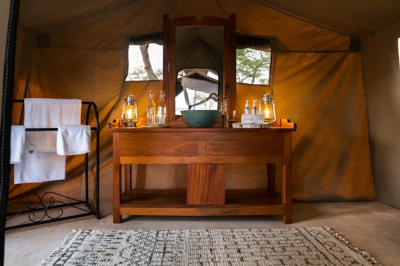 Wooden bathroom vanity with mirror inside a tent, lit by two lanterns; towels on a black rack to the left and a woven rug in front at Honey Bear BUsh Camp, Uganda.