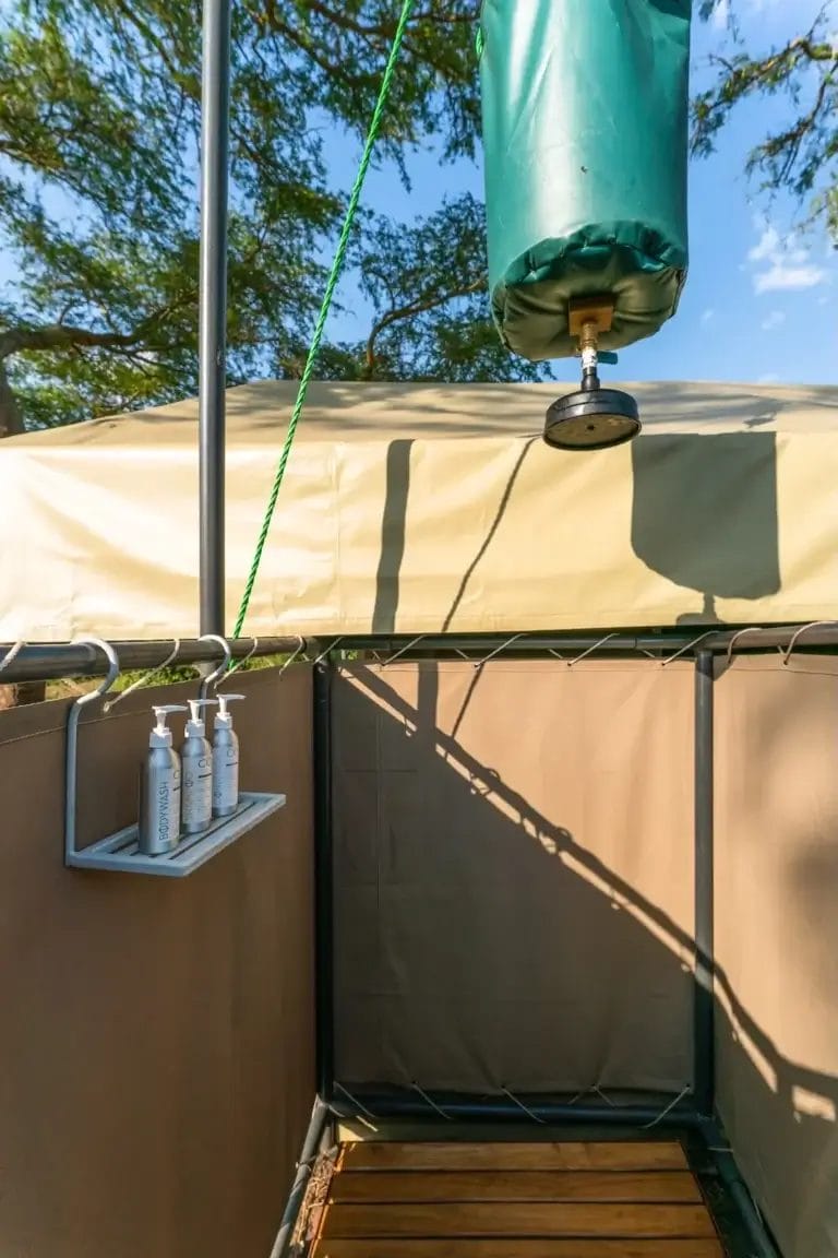Outdoor shower stall with beige privacy walls, wooden platform, and a row of soap bottles on a shelf (head shower hanging above) at Honey Bear Bush Camp, Uganda.