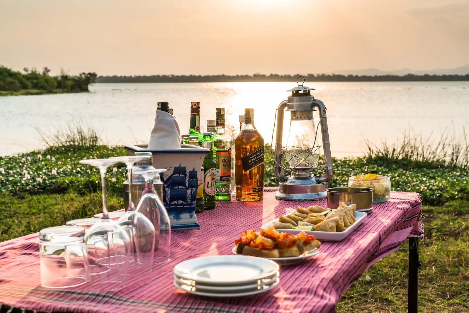 Outdoor picnic table by a lake at sunset with bottles, snacks, a lantern, and a pink tablecloth at Honey Bear Bush Camp,Uganda.