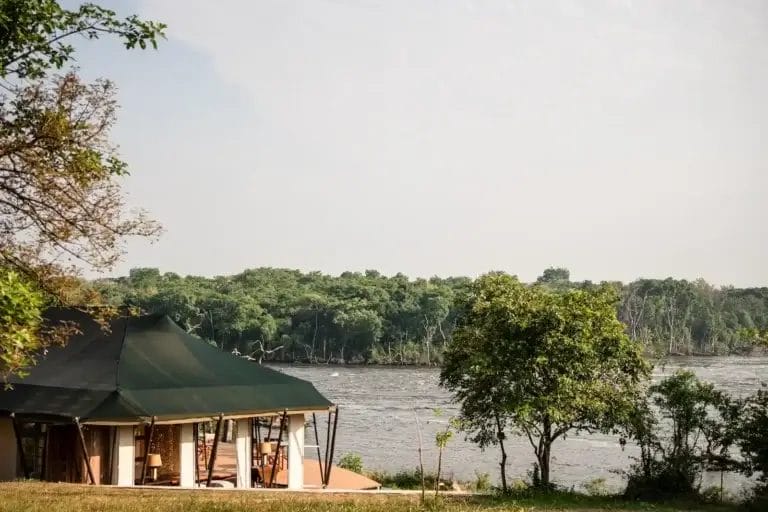 A green canvas tent along the Nile with trees along the shore and a wooden deck visible in the foreground. Kulu Ora Lodge In Uganda - photo credit Felix Studios