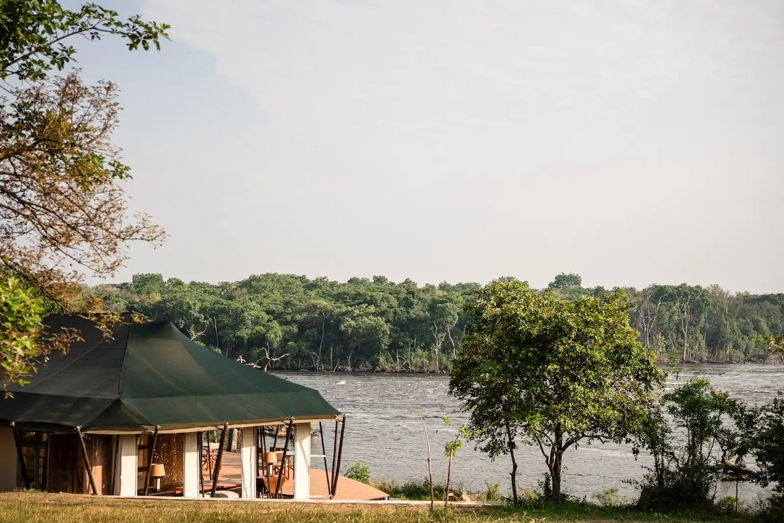 A green canvas tent along the Nile with trees along the shore and a wooden deck visible in the foreground. Kulu Ora Lodge In Uganda - photo credit Felix Studios